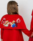 A child wearing a bright red cape-style coat, shown from the back, holding hands with another person in a matching red coat. The back of the coat features a large embroidered floral design wiith two squirrels in white, blue, green, and red.