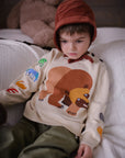 Toddler wearing a soft cotton Brown Bear sweater with embroidered animals on the sleeves, inspired by Eric Carle’s story, sitting on a bed