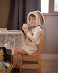 Child wearing Little Goodall embroidered bunny linen jacket and dress, sipping tea while wearing a plush bunny bonnet