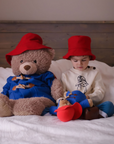 Child and Paddington soft toy sit on a bed wearing matching red bucket hats, with the child playing with plush toys.