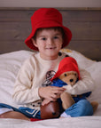 child wearing a red bucket hat and a Paddington Bear sweater sits on a bed, hugging a Paddington Bear plush toy dressed in blue and red.