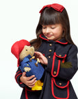 A girl in a dark blue coat holds the Classic Paddington Bear 10" Soft Toy, dressed in a blue toggle coat, red hat, and yellow boots.
