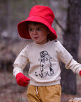 child wears an ivory knit sweater with a black embroidered Paddington Bear design, ribbed cuffs, and button details, pants,a red hat, and gloves,