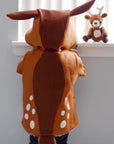 Toddler wears the Frolicsome Fawn Coat, showing its spotted design, long deer tail, and hood with ears. A plush fawn toy sits on the windowsill