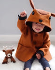 Toddler wearing the Little Goodall Frolicsome Fawn Coat, smiling and raising a hand, with deer hood ears perked up and a plush fawn toy beside them