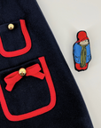 Close-up of a navy blue girls' dress with red-trimmed pockets, gold buttons, and a bow detail, featuring a removable embroidered Paddington Bear