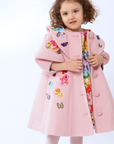 Young girl wearing a pink wool coat with floral embroidery, butterfly appliqués, and a vibrant floral dress underneath