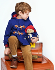 A young child wearing the Little Goodall blue duffle coat, holding a matching Paddington bear, showcasing the cozy hood, wooden toggles, and functional design.