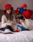 Mother and child wearing matching red wool Paddington bucket hats, sitting on a bed with plush Paddington Bear toys