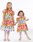 Two girls in matching Rainbow Bloom Party Dresses with puff sleeves and floral butterfly prints, perfect for special occasions and family portraits