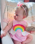 Baby girl wearing Over the Rainbow Bubble Romper in Sunset Pink with a rainbow applique and cloud peplum, a charming summer outfit