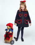 girl wearing a navy wool coat with red trim, gold buttons, and a matching red bow pulls a wicker cart carrying a 16-inch Paddington Bear plush toy