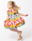 Young girl twirling in the Rainbow Bloom Dress, featuring a vibrant floral and butterfly print, puff sleeves, and a tie waist.