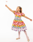 Girl in Rainbow Bloom Party Dress, holding a butterfly wand. Vibrant floral print with puff sleeves and a tie waist, perfect for special occasions