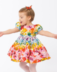 Girl twirling in a vibrant Rainbow Bloom Party Dress with puff sleeves, a pink waist bow, and a floral butterfly print, perfect for special occasions