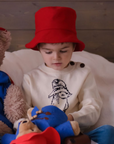 Child wearing a red wool Paddington bucket hat, sitting on a bed with Paddington Bear toys