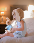 toddler wearing Scooter Bunny Linen Jon Jon sits on a bed with a plush bunny, showcasing the embroidered vintage-style outfit