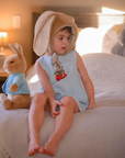 Toddler wearing light blue linen romper with embroidered scooter bunny, sitting on a bed with a plush bunny. Heirloom-quality baby clothing