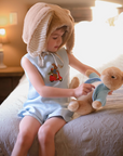Toddler wearing a light blue linen romper with embroidered scooter bunny, sitting on a bed with a plush bunny. Heirloom-quality baby outfit