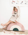 kid wearing a Snowshoe Rabbit Coat with bunny ears, toggle fasteners, and Merino wool fabric, sitting on a pastel rainbow cushion.