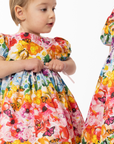 Toddler wearing Rainbow Bloom Dress with vibrant floral and butterfly print. Puff sleeves and a waist tie add charm to this colorful outfit