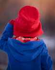 Back view of a toddler wearing a red hat and blue coat, standing outdoors in a winter