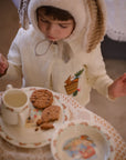 Toddler in embroidered Little Goodall linen bunny jacket enjoying tea time. Heirloom-quality spring cardigan with bunny and bushel basket design.