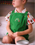Toddler boy wearing the Very Hungry Caterpillar™ Party Food Shirt, a short-sleeve button-up with a colorful food print, paired with green overall