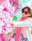 Girl wearing Yolanda and Tatty pink wool coat with zebra appliqués, styled with a skirt and bow, in a floral setting