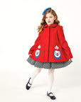 Girl modeling red coat while wearing a polkadot dress underneath and a blue bow on top of her head.