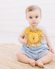 Baby wearing Blue Gingham Lion Romper for boys and girls with embroidered lion face and ruffled mane, sitting on a woven rug, looking at the camera