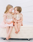 Two children smiling in coordinating Little Goodall bunny outfits—a pink gingham Bunny Love Dress and Bunny Face Romper with floppy ears and carrot details