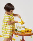 A child in a lemon-print dress squeezes a lemon into a glass pitcher on a wooden table with fresh lemons and limes.