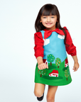Adorable toddler in a red, blue, and green jumper dress with cloud and landscape motifs, perfect for imaginative play.