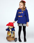 girl in a blue wool duffle coat with wooden toggles, standing beside a wicker cart carrying a Paddington Bear plush toy in a matching coat and red hat
