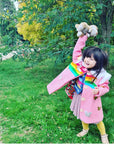 Smiling girl in a pink wool coat with rainbow appliqués and butterflies, playing outside with a stuffed bunny, coat lining peeking through