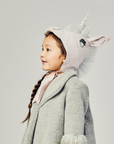 Young girl wearing a pale pink wool unicorn hat with a silver mane and satin ribbon ties, styled with a cozy grey winter coat