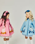 Two girls wearing blue and pink wool coats with embroidered dog designs, tassels, and coordinating bows, featuring Willa Heart artwork.