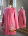 Pink wool A-line coat on form, featuring embroidered dog motifs, Peter Pan collar, and hidden placket, with back view reflected in mirror