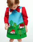 Child wearing the Little Cloud jumper dress with a red bow blouse, featuring appliqué houses, trees, and clouds inspired by Little Cloud by Eric Carle