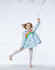 Girl in a sky blue wool coat with rainbow and butterfly details, holding white balloons while posing playfully