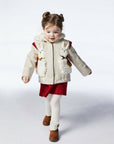 A young girl wearing the Lucky Llama Coat, a merino wool jacket with faux fur lining, embroidered llama details, and a cozy hood with a red tassel.