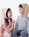 children wearing cozy beige hats with blue linings; the design features playful bunny-inspired details, made from 100% Merino wool and fully lined with soft fleece for warmth.