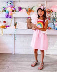 Girl in pink rainbow sundress holding cotton candy in a toy-filled boutique, wearing strappy sandals and a unicorn headband