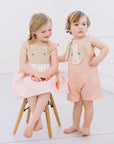 Two children wearing Little Goodall bunny-themed outfits—pink gingham Bunny Love Dress with carrot details and coordinating pink Bunny Face Romper
