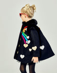Girls' Queen of Hearts coat in black wool with rainbow stripes, metallic heart appliqués, and faux fur collar, shown from a side angle.