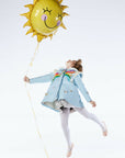 A girl in the Rainbow Butterfly wool coat, white tights, and ballet slippers leaps joyfully, holding a yellow sun balloon