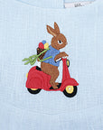 Close-up of embroidered bunny riding a red scooter on light blue linen romper. Detailed heirloom-quality stitching.