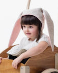 A child dressed in white, wearing Little Goodall's grey Snowshoe Rabbit Bunny Hat with soft floppy ears, sitting in a wooden wagon