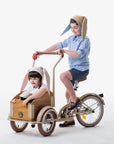 Two children on a tricycle, with one wearing the Snowshoe Rabbit Bunny Hat, grey with floppy ears